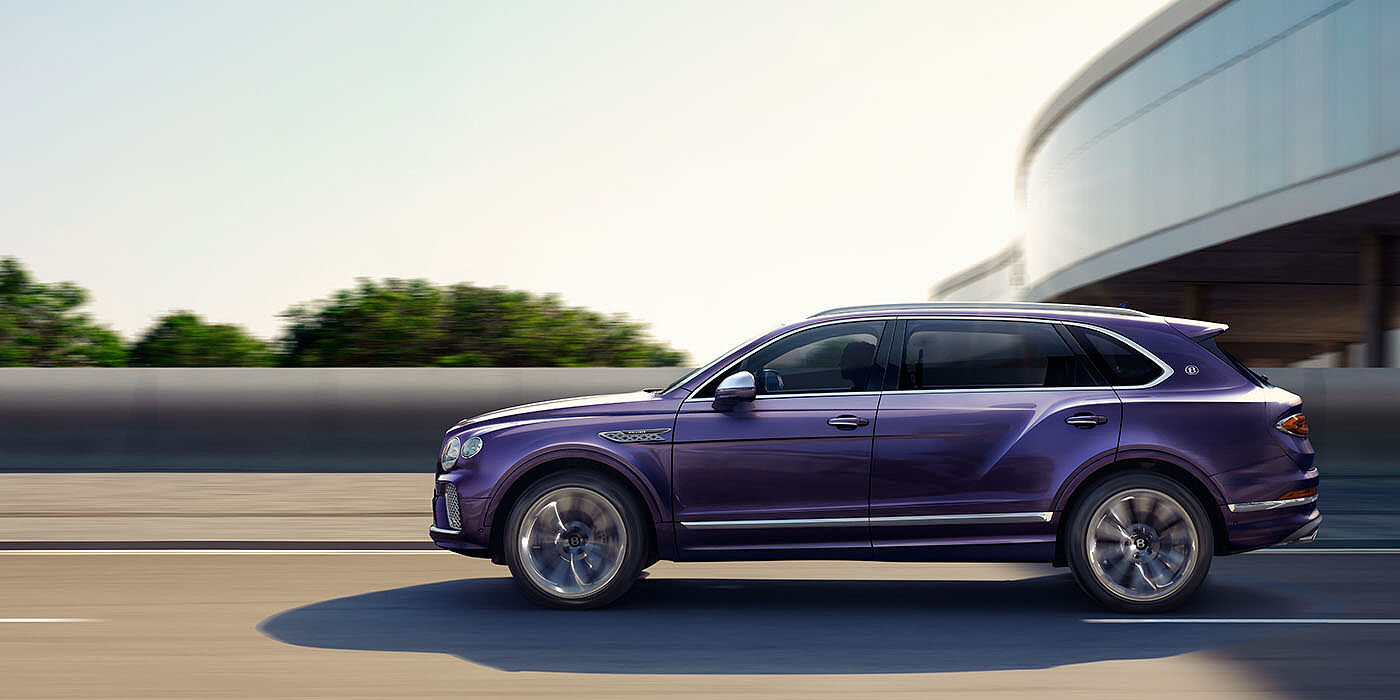 Bentley Birmingham Bentley Bentayga Extended Wheelbase Mulliner luxury SUV profile in Tanzanite Purple paint, driving on an country road past trees.