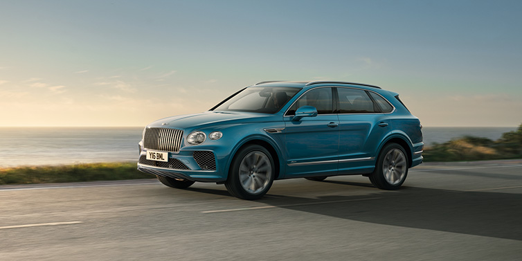 Bentley Birmingham Bentley Bentayga Azure SUV in Topaz blue paint driving dynamically by the ocean with 22 inch 10 spoke directional wheels