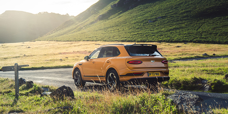 Bentley Birmingham Bentley Bentayga S SUV rear three quarter in Sunburst Gold paint on a road surrounded by green hills