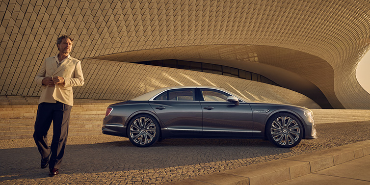 Bentley Birmingham Bentley Flying Spur Mulliner sedan in Rose Gold over Magnetic duotone paint parked by a curved wall with person in suit jacket standing on the left